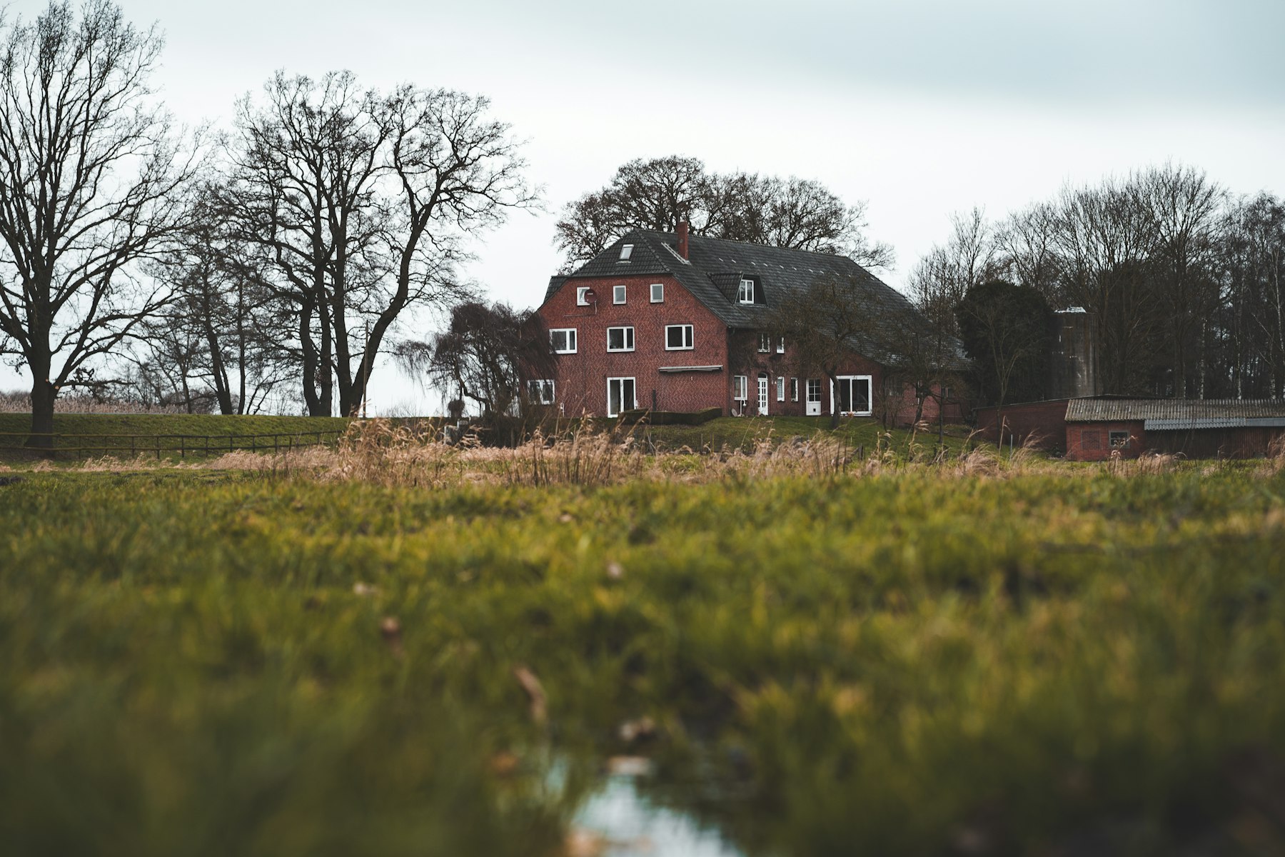 Rotes Backsteinhaus mit Walmdach hinter einer Wiese und kahlen Bäumen – geerbte Immobilie verkaufen - Quelle: Unsplash (Lizenz: Unsplash License)