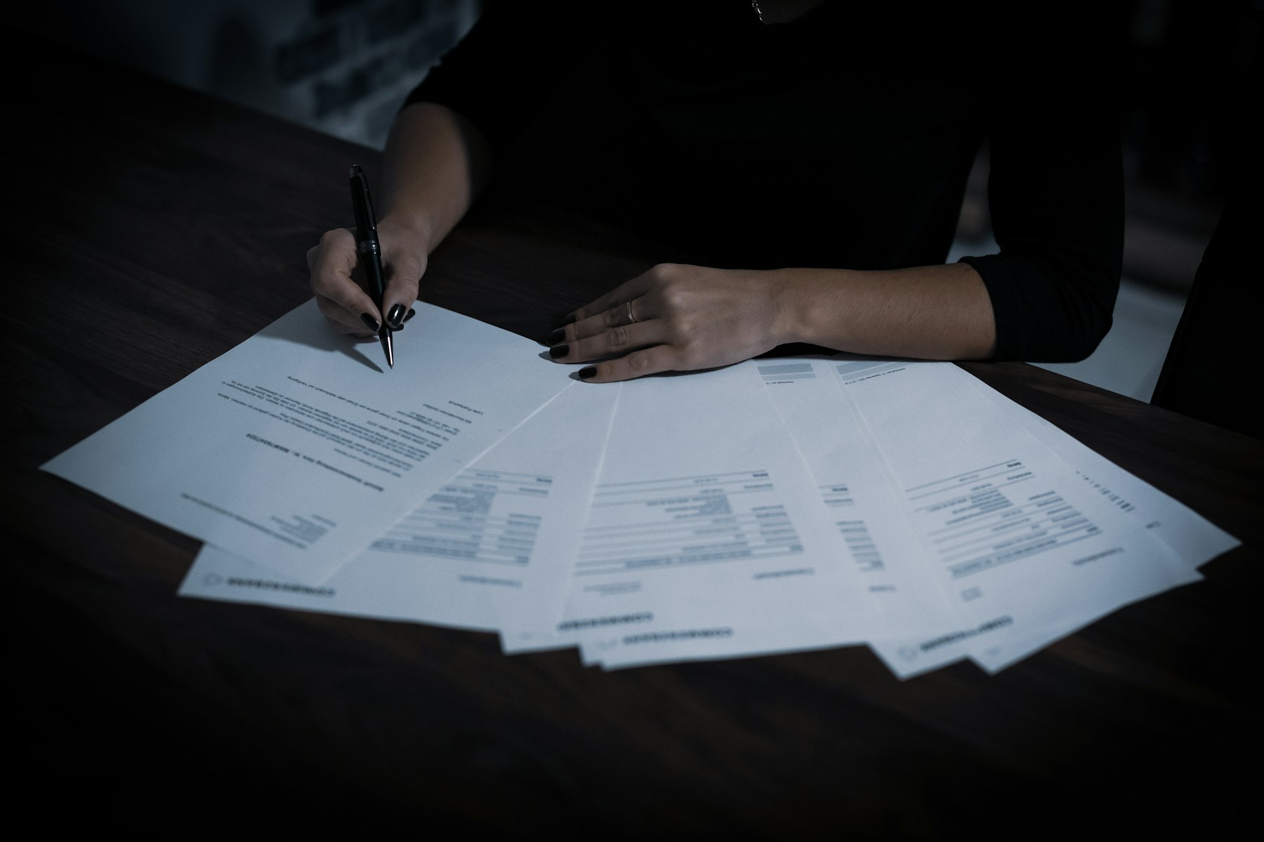 Person unterschreibt an einem dunklen Tisch mehrere ausgebreitete Dokumente – Vertraulichkeitsvereinbarung beim diskreten Verkauf - Quelle: Unsplash (Lizenz: Unsplash License)